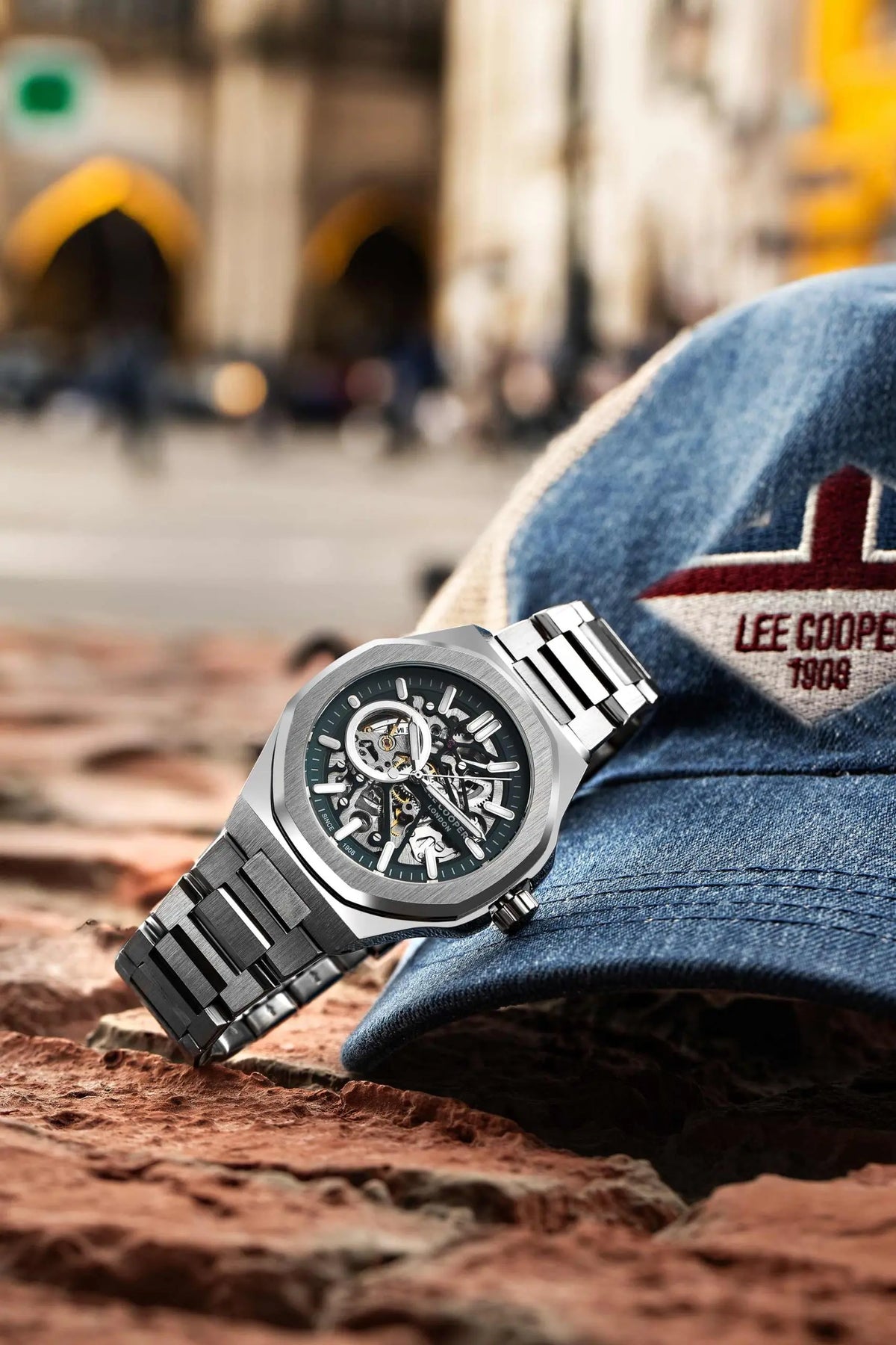 A luxury stainless steel skeleton wristwatch with a black dial and integrated metal bracelet, resting on a textured red-brown stone surface next to a blue denim cap with 'LEE COOPER 1909' embroidery, with a blurred outdoor city street and historic building in the background.