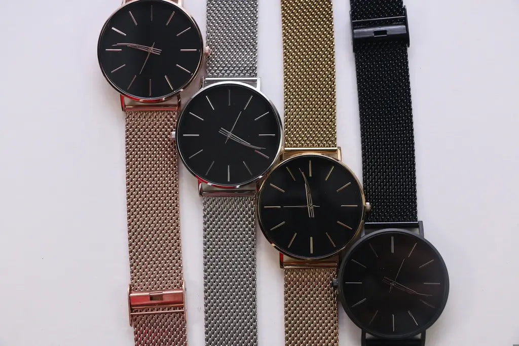 Four minimalist analog wristwatches with round black dials and mesh metal bands in rose gold, silver, gold, and black, arranged vertically on a plain white background.