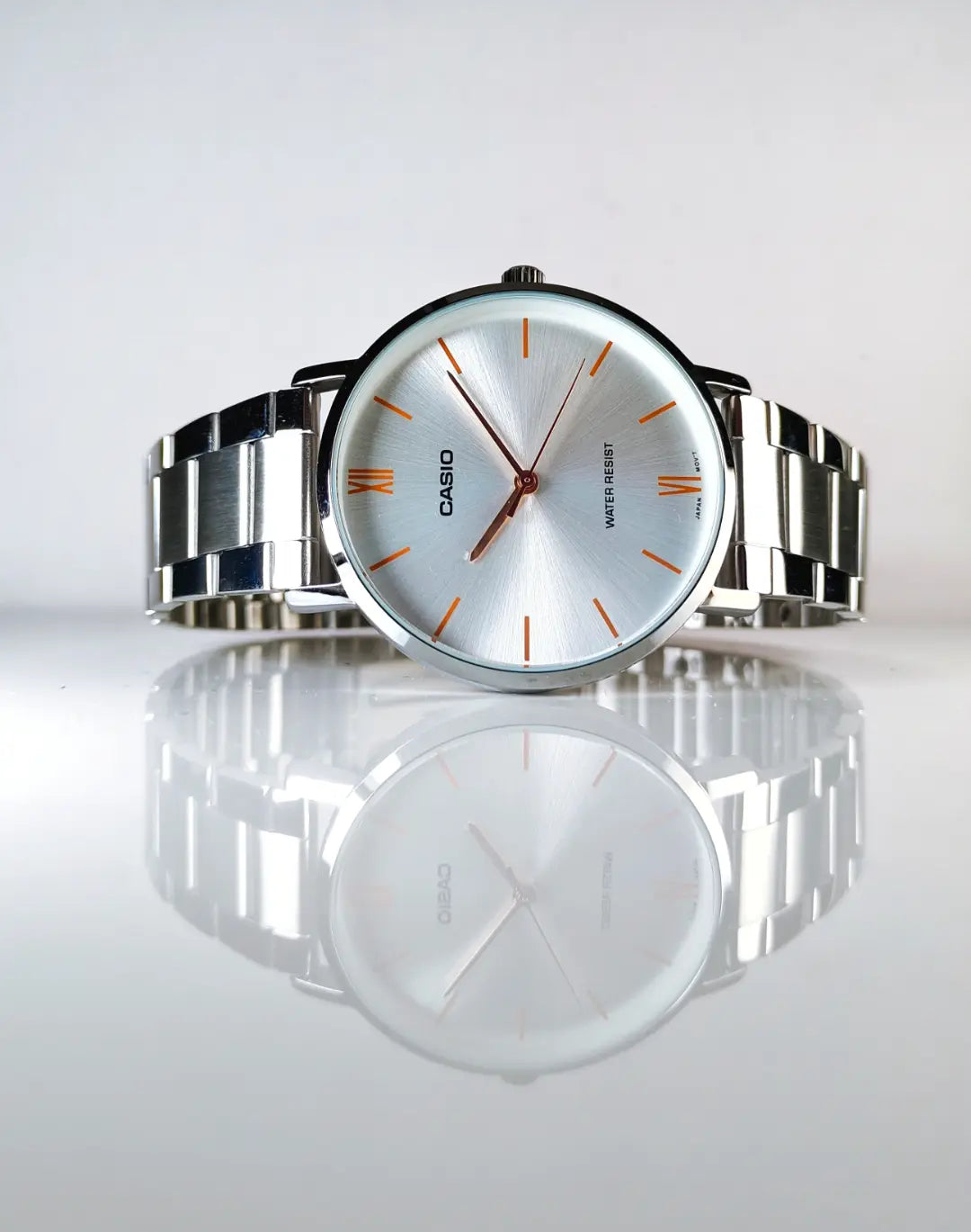 A Casio analog wristwatch with a silver stainless steel link bracelet, a silver dial, and orange accents on the hands and hour markers, displayed on a reflective white surface.