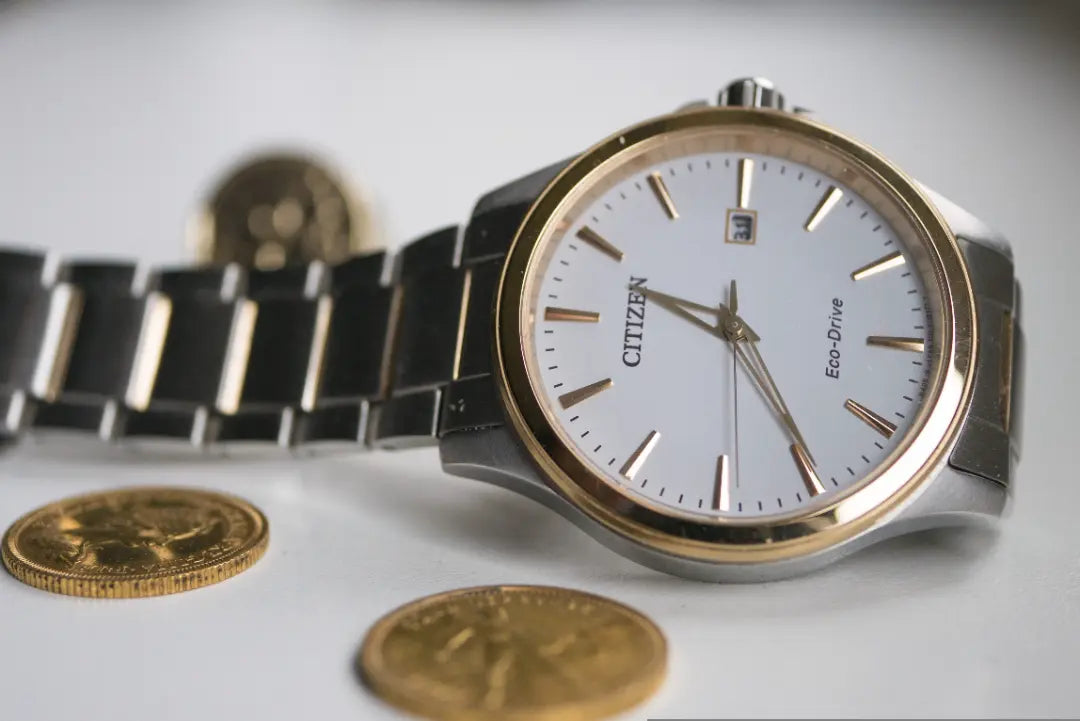 Citizen Eco-Drive watch with a silver and gold-tone metal bracelet, a gold bezel, and a white dial, styled with gold coins on a light gray background.