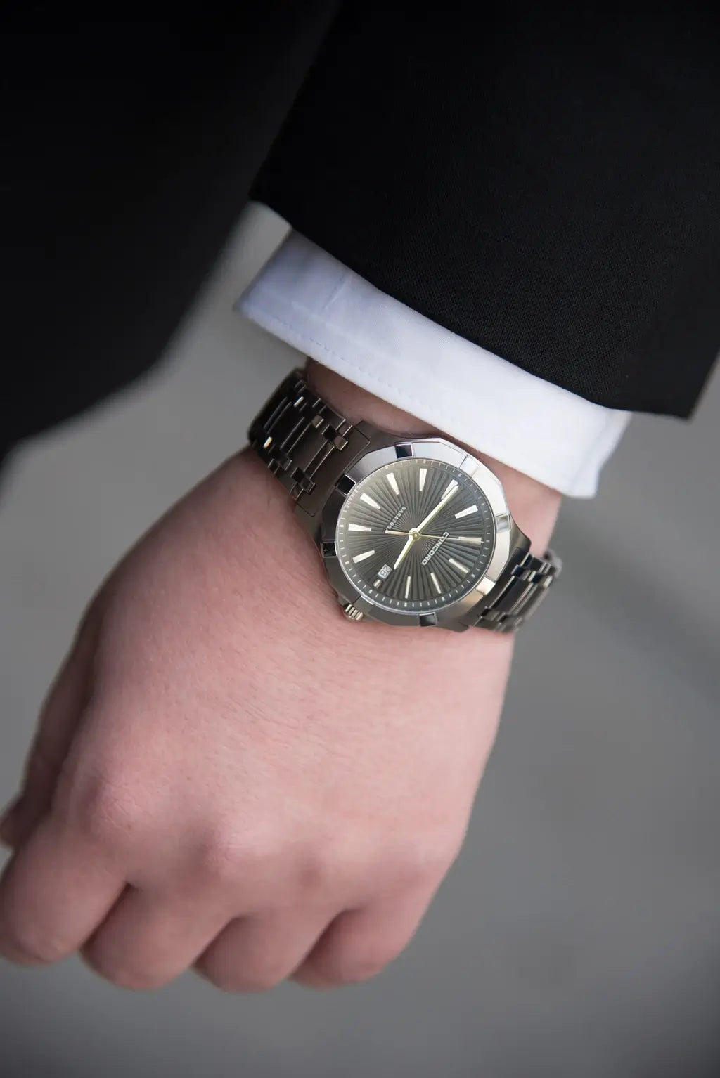 A close-up of a silver-toned metal wristwatch with a dark dial and matching metal link bracelet, worn on a person's wrist with a white dress shirt cuff and black suit jacket visible.