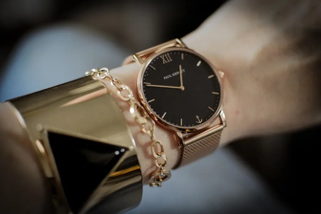 A close-up lifestyle shot of a Paul Weller wristwatch with a black dial and rose gold case, worn on a wrist alongside a gold chain bracelet and a chunky gold cuff bracelet.